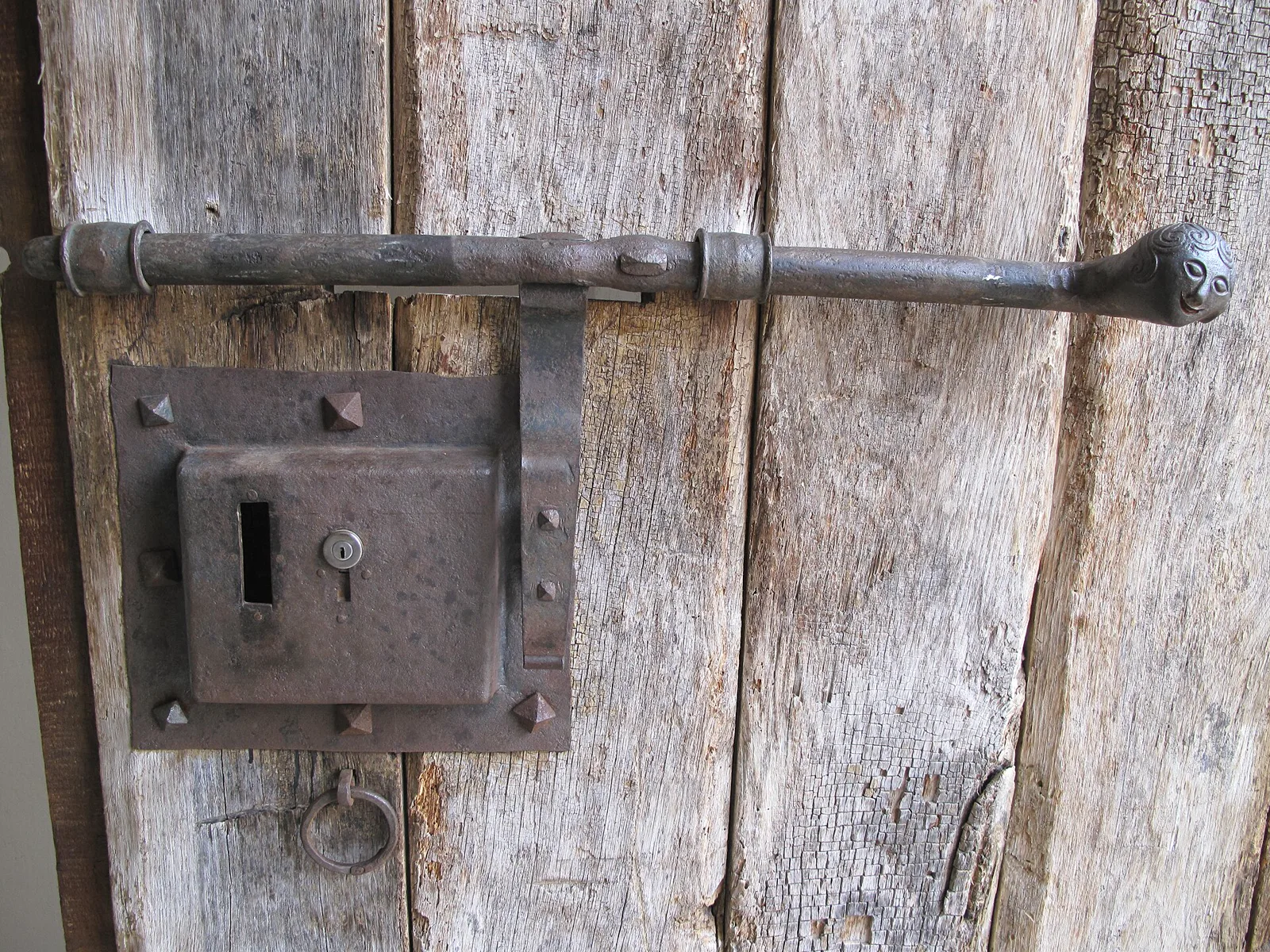 A rusted iron box lock and a long iron bolt with a carved knob fixed to the weathered gray planks of an old wooden church door, with an iron ring below