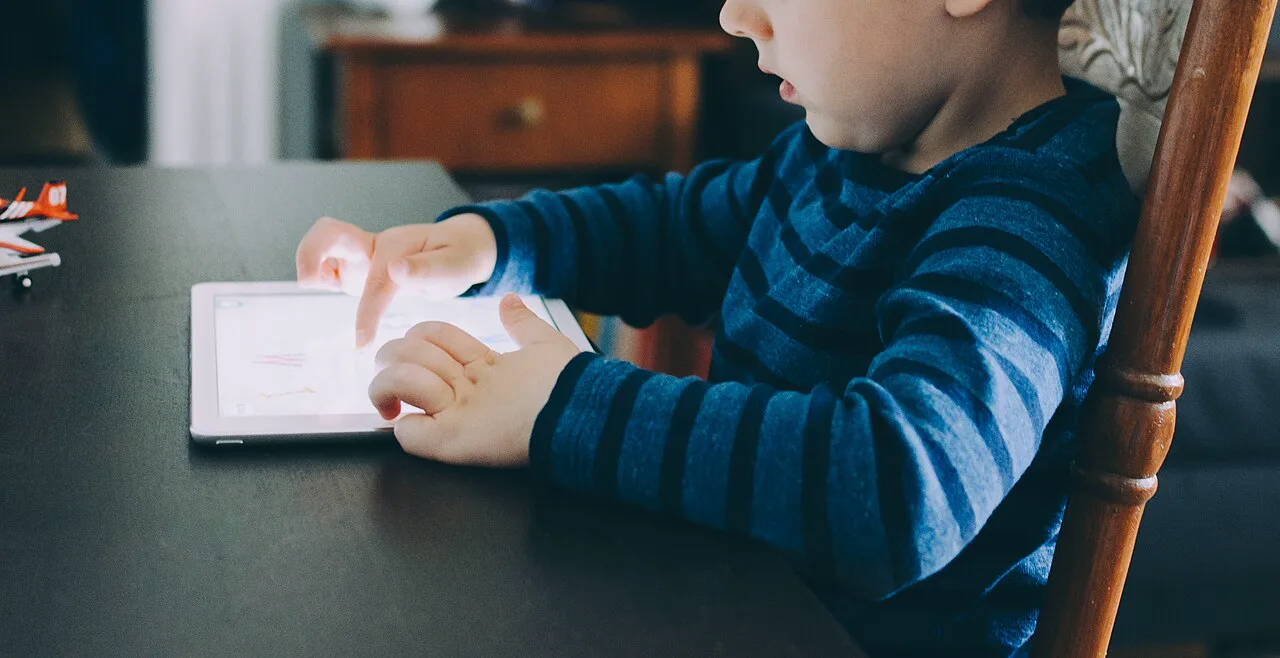 A small child in a striped blue sweater, seen from the side with the eyes out of frame, taps a tablet lying on a dark table while sitting on a wooden chair