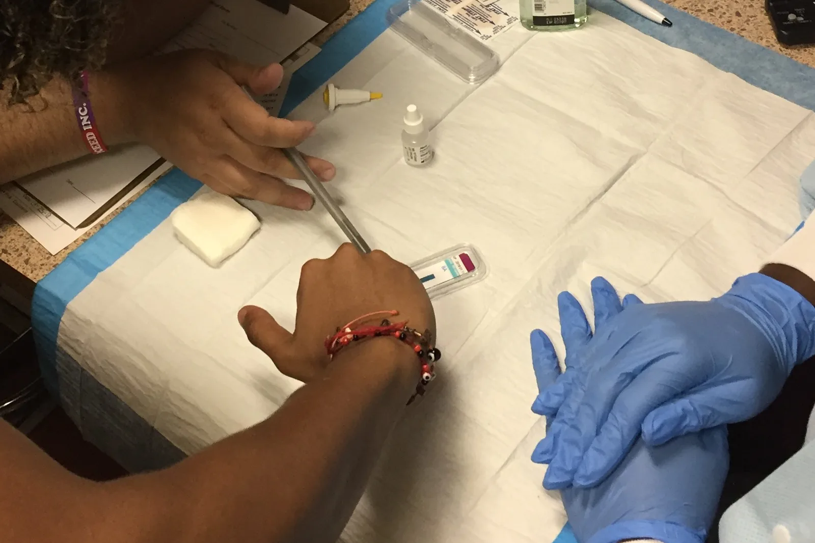 Close view of hands over a paper-covered table in a mobile testing van: one person holds a lancet near a rapid test cassette and a small bottle, while a counselor in blue gloves rests her hands on the table