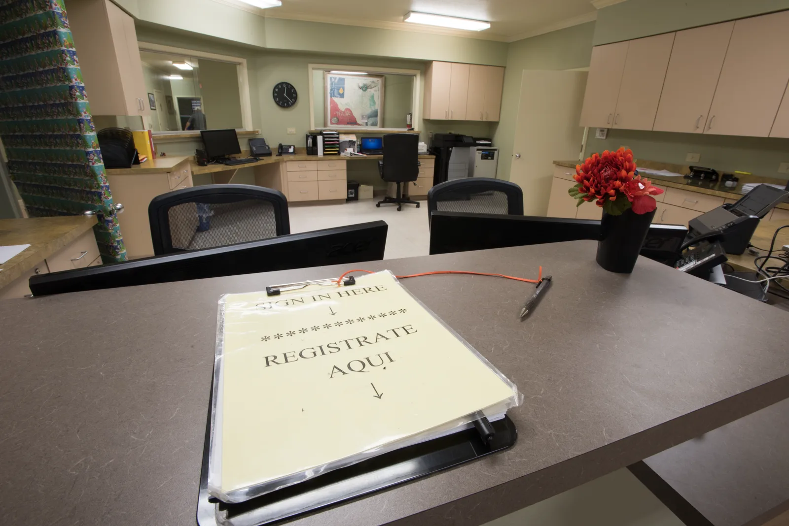 A clipboard on a reception counter reads Sign in here and Registrate aqui, with empty office chairs, desks and a vase of red flowers behind it