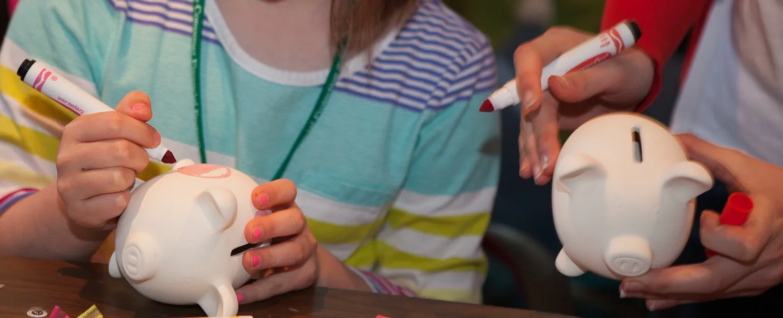 Children's hands holding white ceramic piggy banks and red markers over a wooden table, one child decorating a piggy bank and another holding one up