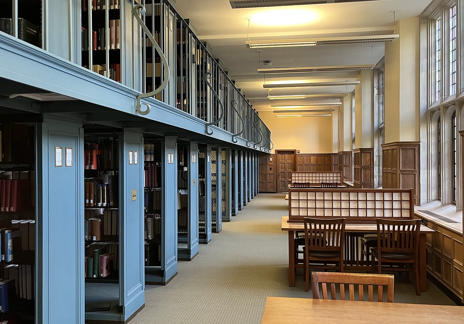 An empty library reading room with pale blue book stacks and a railed upper gallery on the left, wooden study tables and chairs on the right and tall windows along the wall