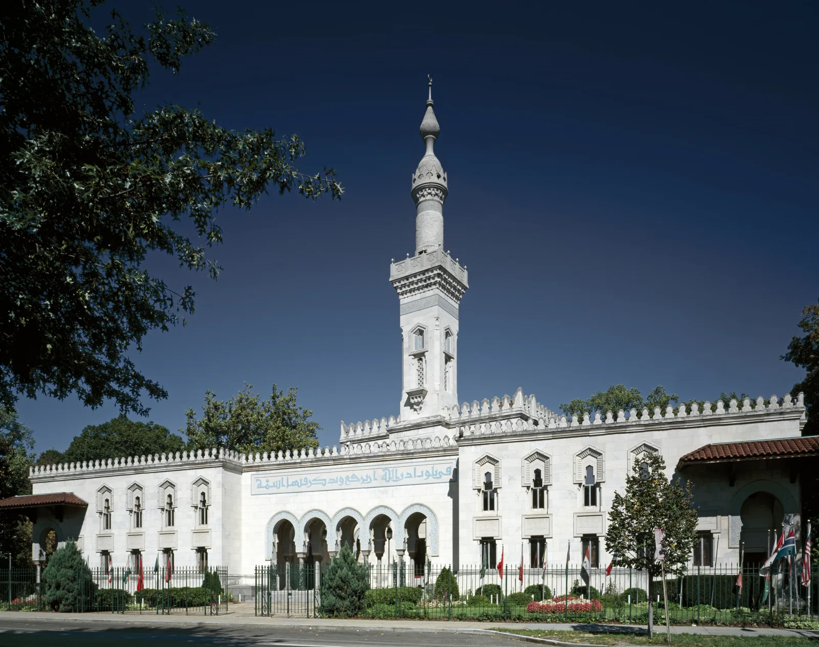 A white stone mosque with a tall minaret, arched entrance and a band of blue calligraphy, behind a low iron fence and gates, under a deep blue sky with trees at the left