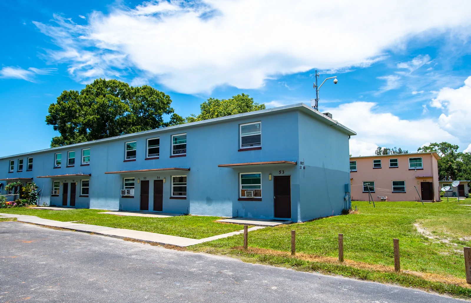Two-story pale blue apartment buildings with brown doors and window air conditioners, set on a lawn beside a paved street under a blue sky with large trees behind them