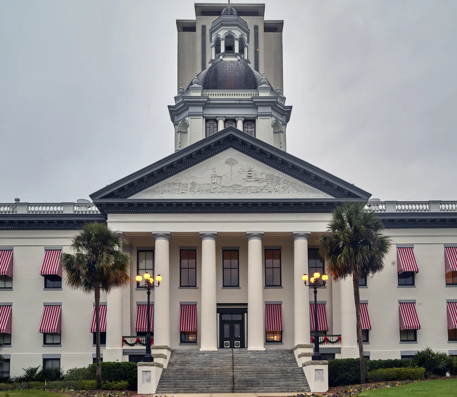A white columned building with red and white striped awnings, palm trees and a domed cupola, with a tall modern tower rising behind it under an overcast sky