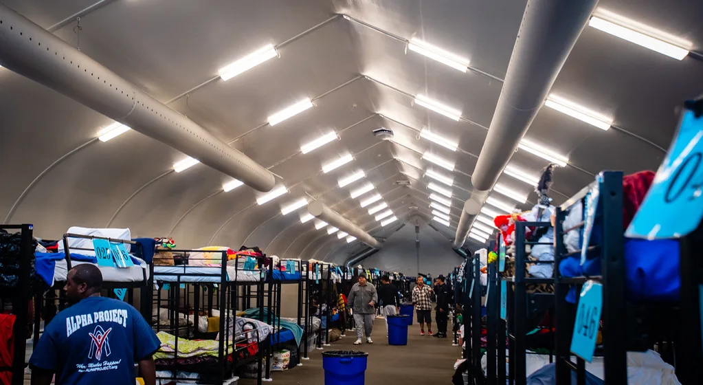 Rows of metal bunk beds with blankets and numbered tags line both sides of an aisle under a long white arched tent roof with ventilation ducts and bright ceiling lights, a few people standing far down the aisle