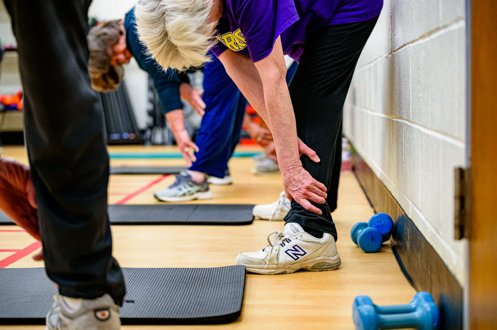 Older adults in an exercise class bend forward to stretch beside floor mats in a gym, faces turned down, with blue hand weights by the wall
