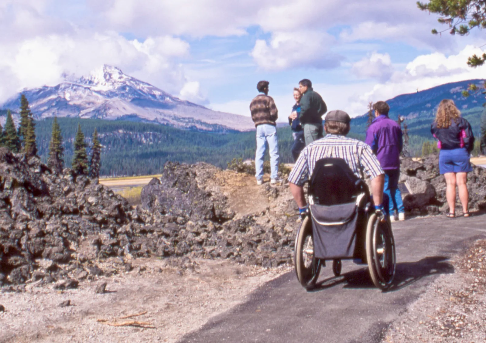 A man in a wheelchair, seen from behind, rolls along a paved trail past black lava rock toward a group of visitors, with a snow-capped mountain in the distance