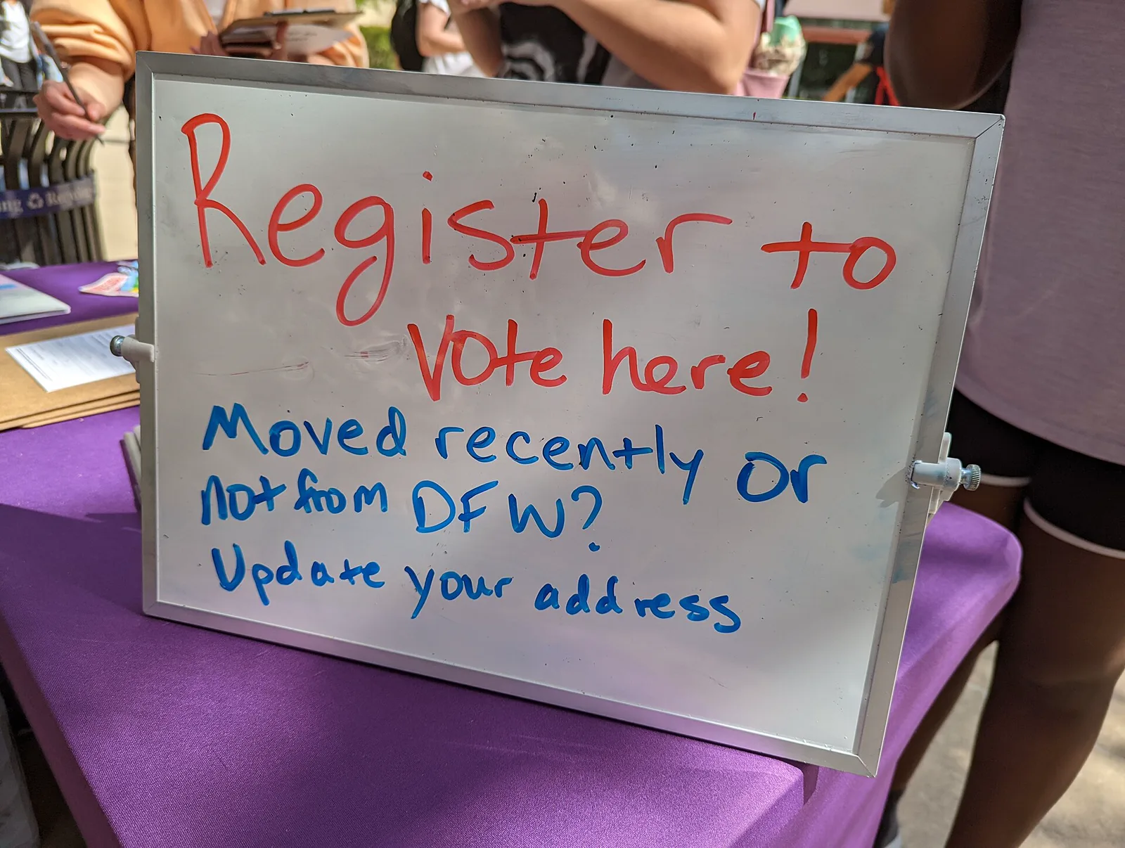 A handwritten whiteboard sign reading Register to vote here! Moved recently or not from DFW? Update your address, propped on a table with a purple cloth and clipboards, with people standing behind it, their faces out of frame