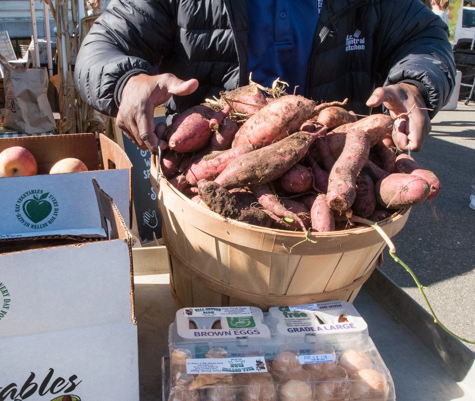 A man in a dark jacket, his face out of the frame, holds both hands over a wooden bushel basket full of freshly dug sweet potatoes, beside cartons of eggs and a box of apples