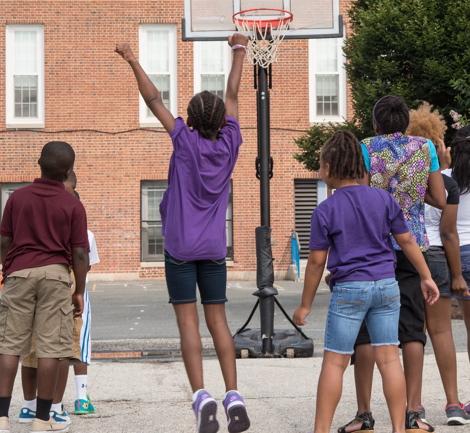 Children seen from behind on a schoolyard basketball court, one girl in a purple T-shirt jumping toward the hoop in front of a red brick school building