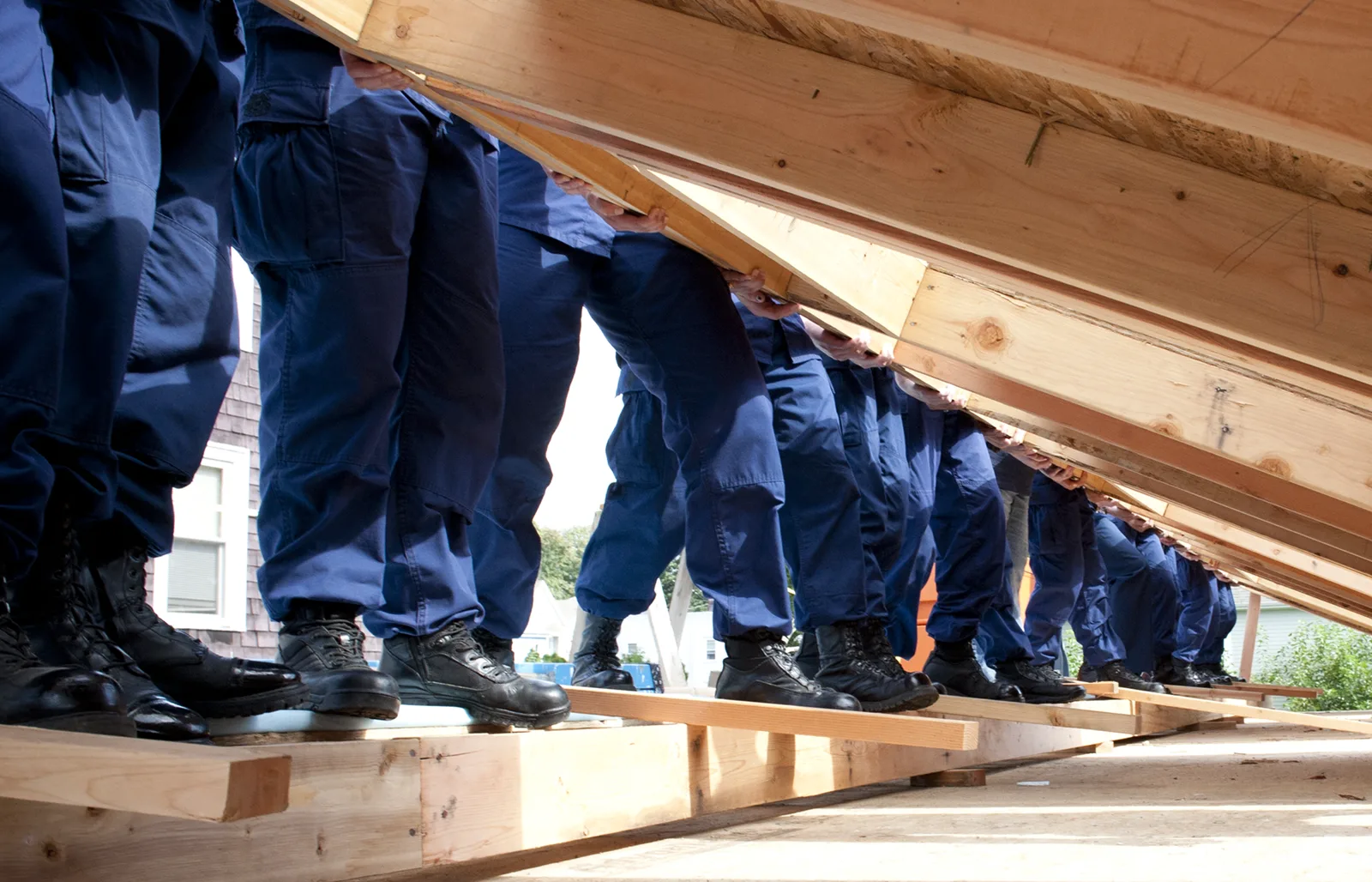 A row of people in navy work uniforms and black boots, seen from the knees down, push a framed wooden wall upright on a plywood house floor
