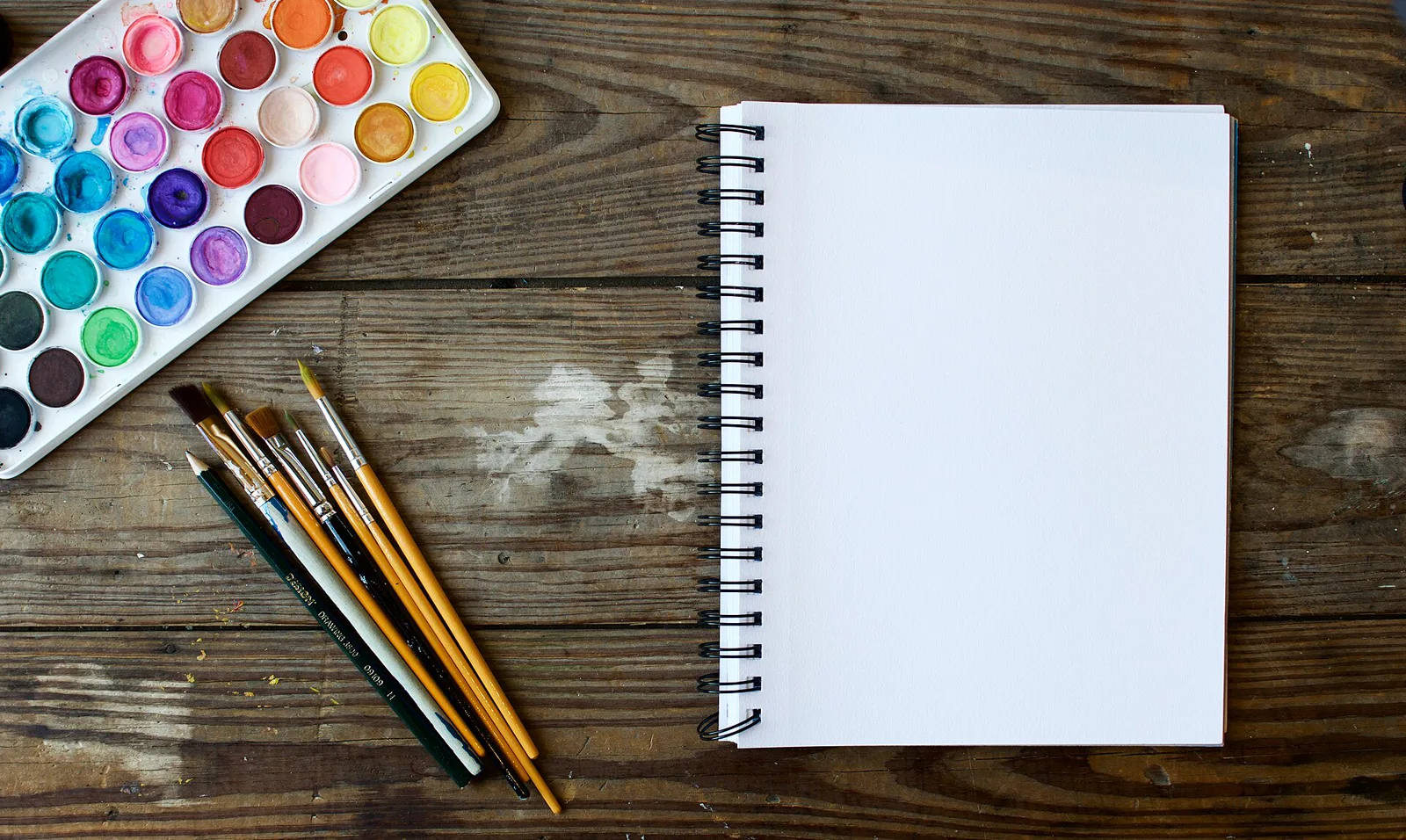 A watercolor paint set, a row of brushes and a pencil beside a blank spiral sketchbook on a weathered wooden table