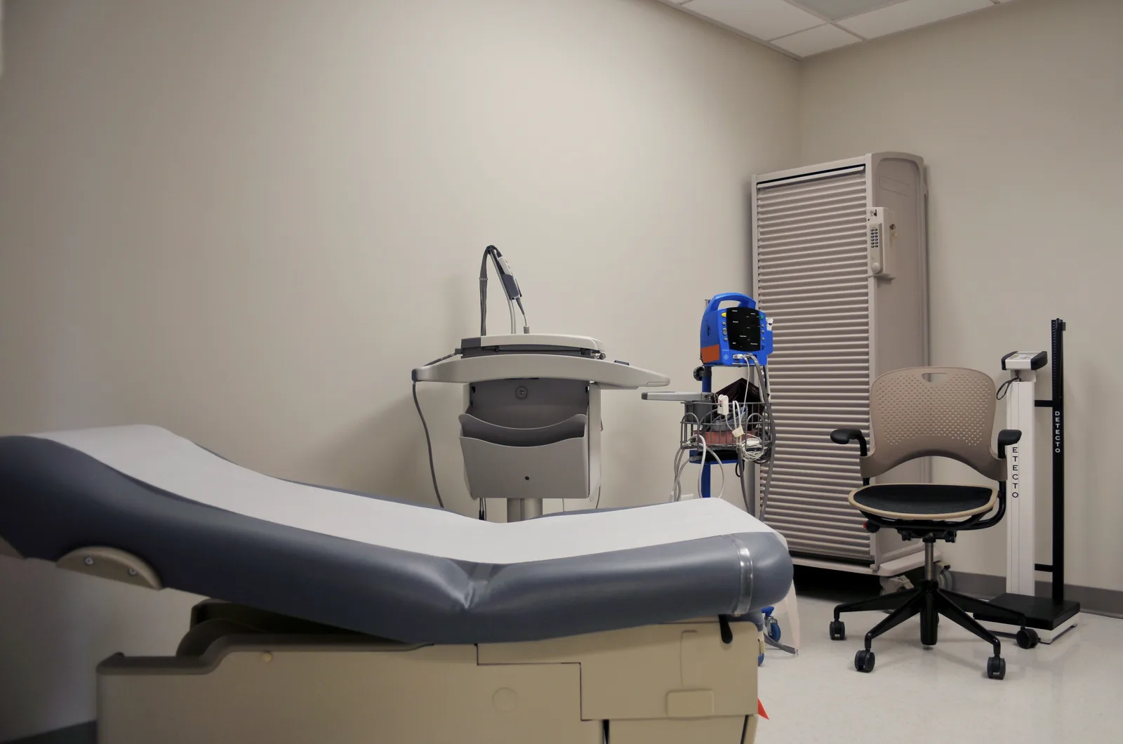 An empty medical examination room with a padded exam table in the foreground, a wall cart, a blue vital signs monitor on a stand, a rolling chair, a standing scale and a tall supply cabinet