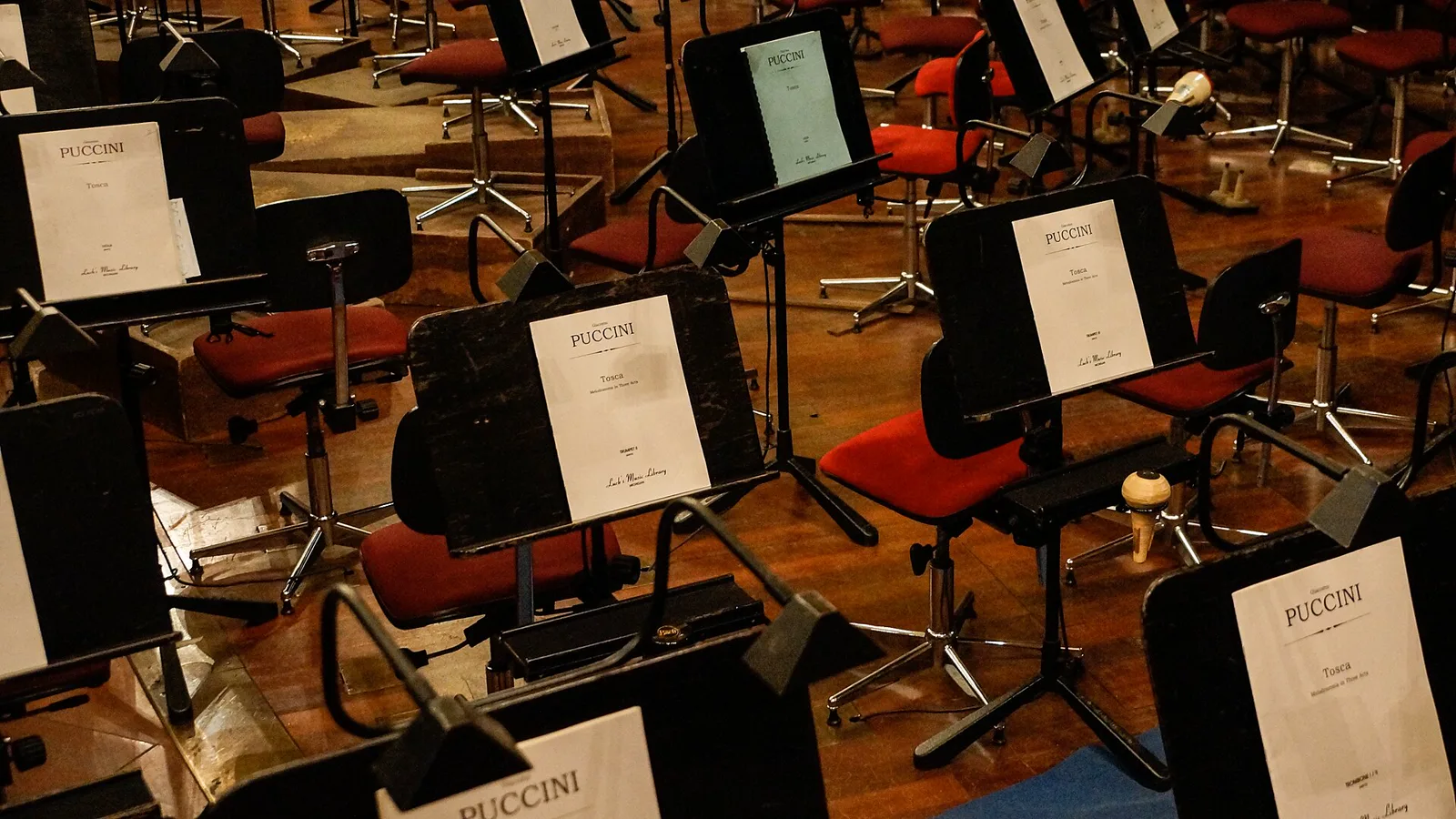 Empty red chairs and black music stands holding parts of Puccini's Tosca in an orchestra pit, with a trumpet mute resting on a stand
