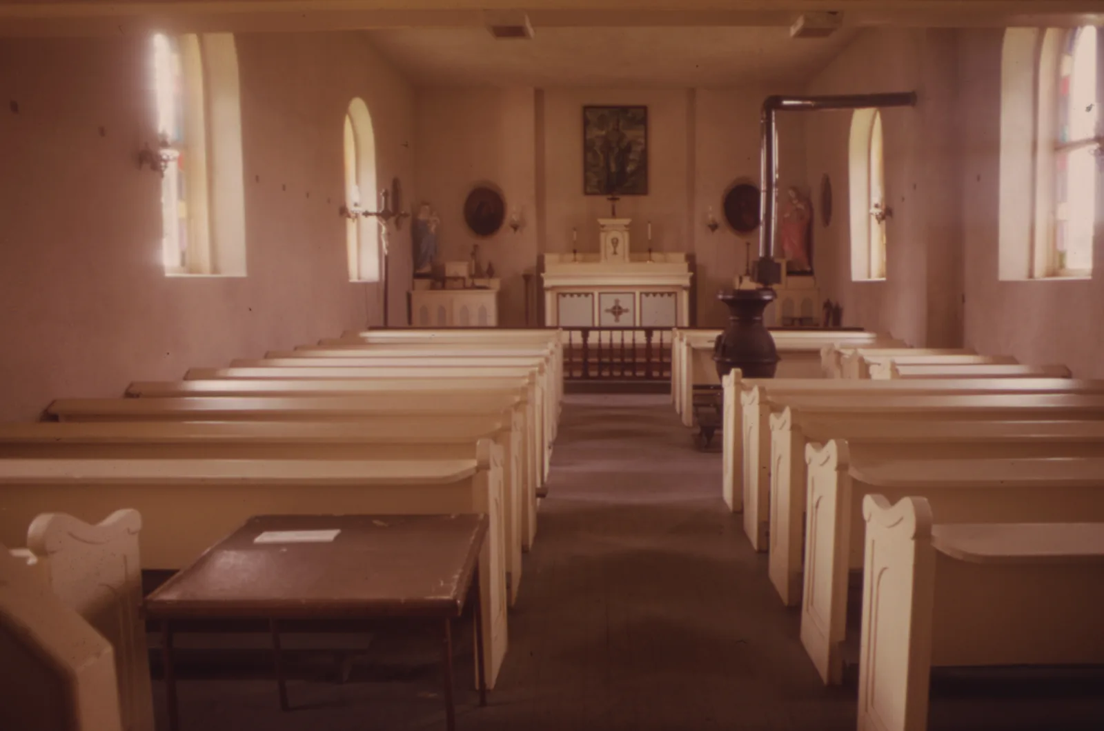 The empty interior of a small church with rows of cream painted wooden pews, a cast iron wood stove in the aisle, arched windows and a plain white altar