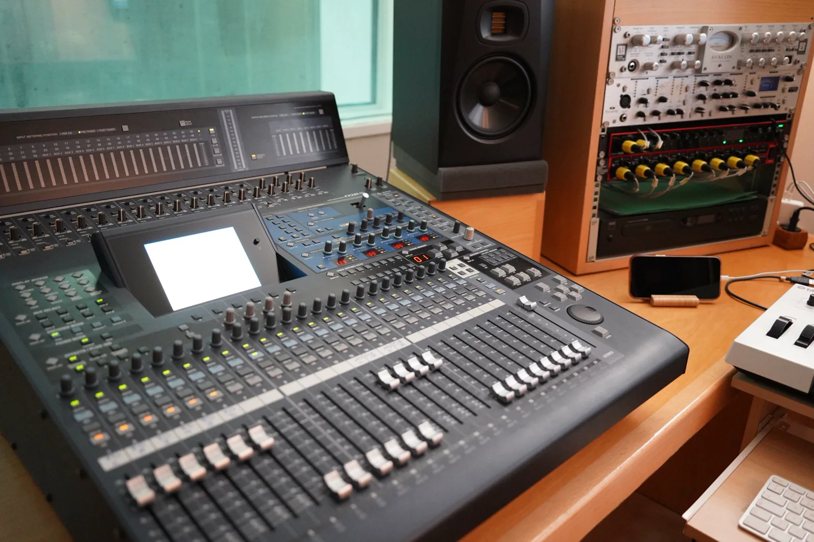 A large digital audio mixing console with rows of faders, knobs and a small screen sits on a wooden desk, with a studio monitor speaker and a rack of audio equipment behind it