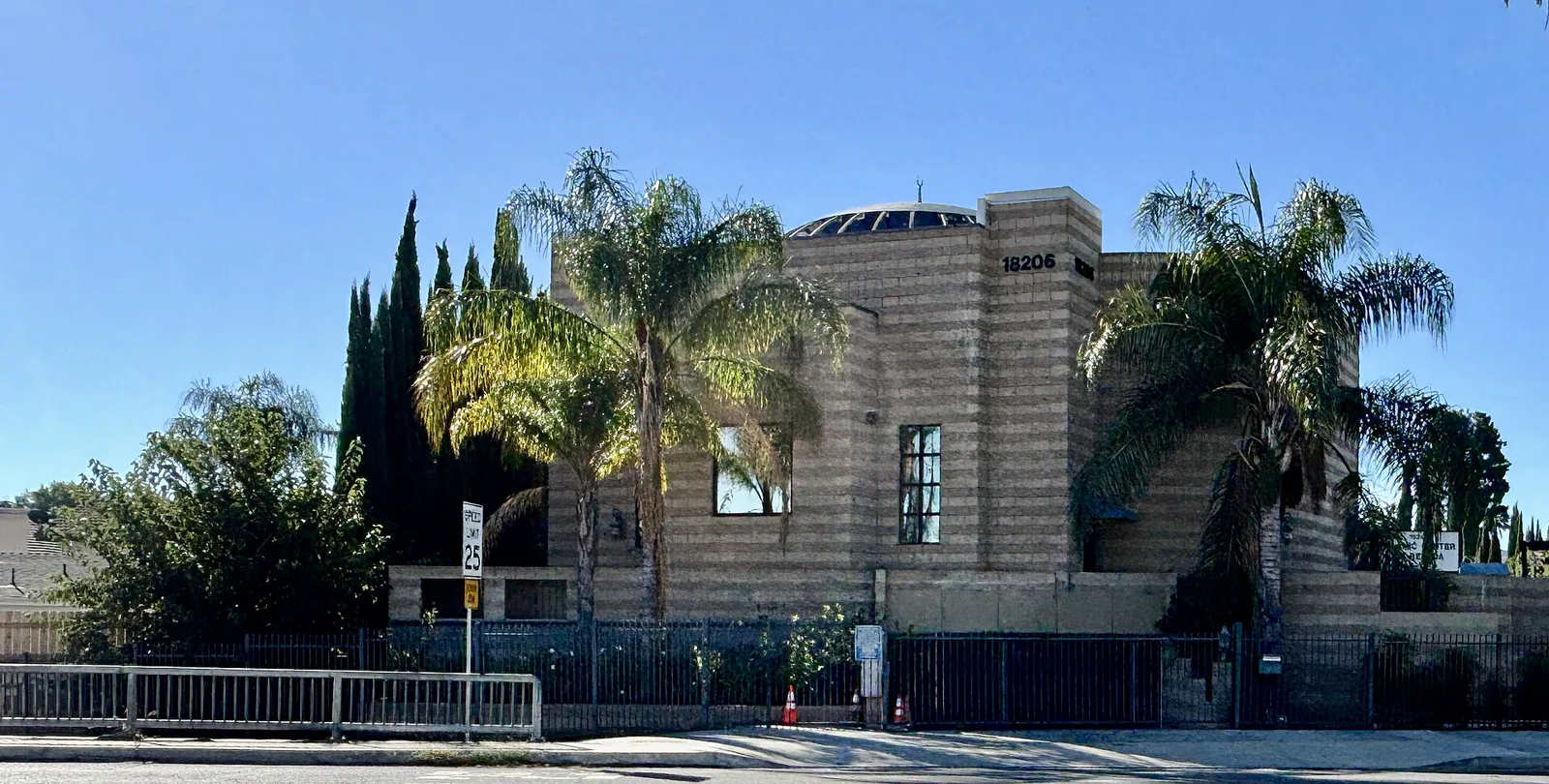 A tan block mosque with a small dome and a crescent on top, behind palm trees and a tall black iron fence with closed gates along the sidewalk