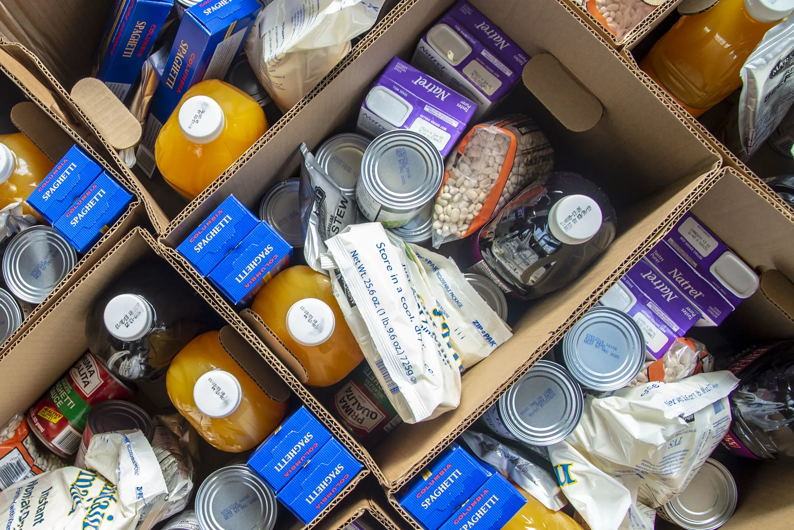 Open cardboard boxes seen from above, packed with boxes of spaghetti, bottles of juice, canned goods, bags of dried beans and cartons of shelf-stable milk