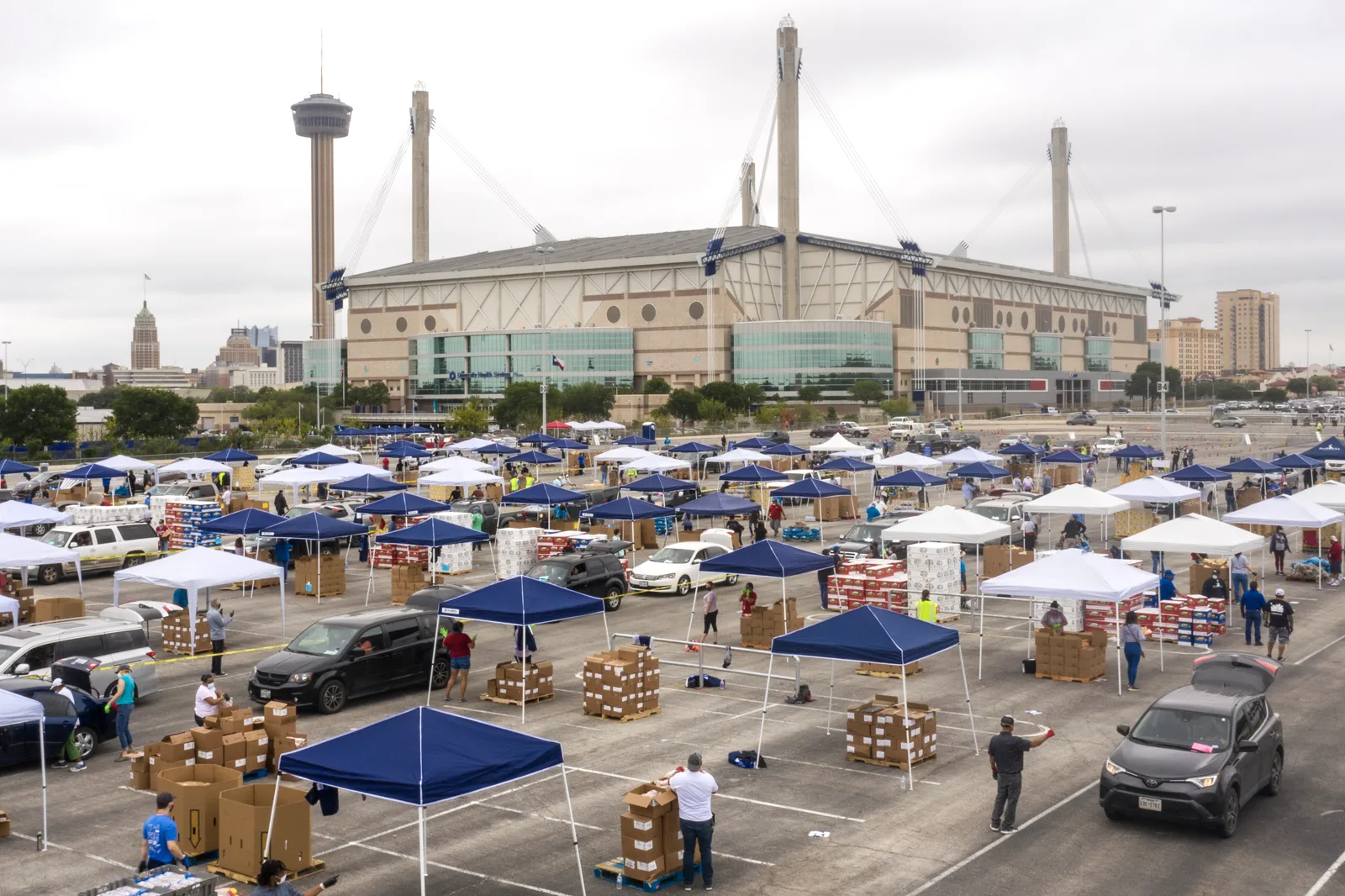 Rows of blue and white canopy tents, pallets of cardboard boxes and cars in a large parking lot, with volunteers loading food, a domed stadium with tall white masts and an observation tower behind under a cloudy sky