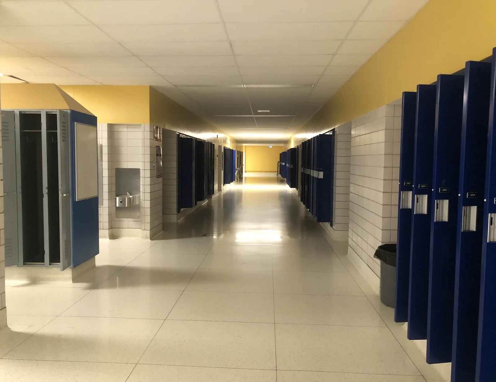 An empty school hallway with white tile walls and a yellow band near the ceiling, lined with blue lockers whose doors stand open