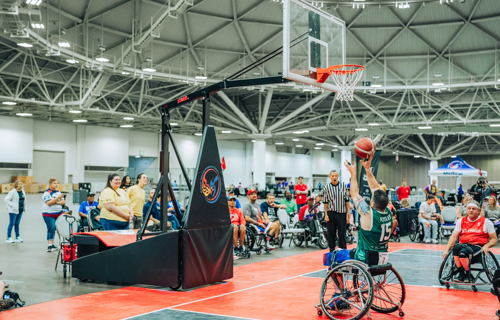 A player in a green jersey, seen from behind in a sports wheelchair, shoots a basketball at a hoop in a large convention hall as a referee, players in wheelchairs and spectators look on