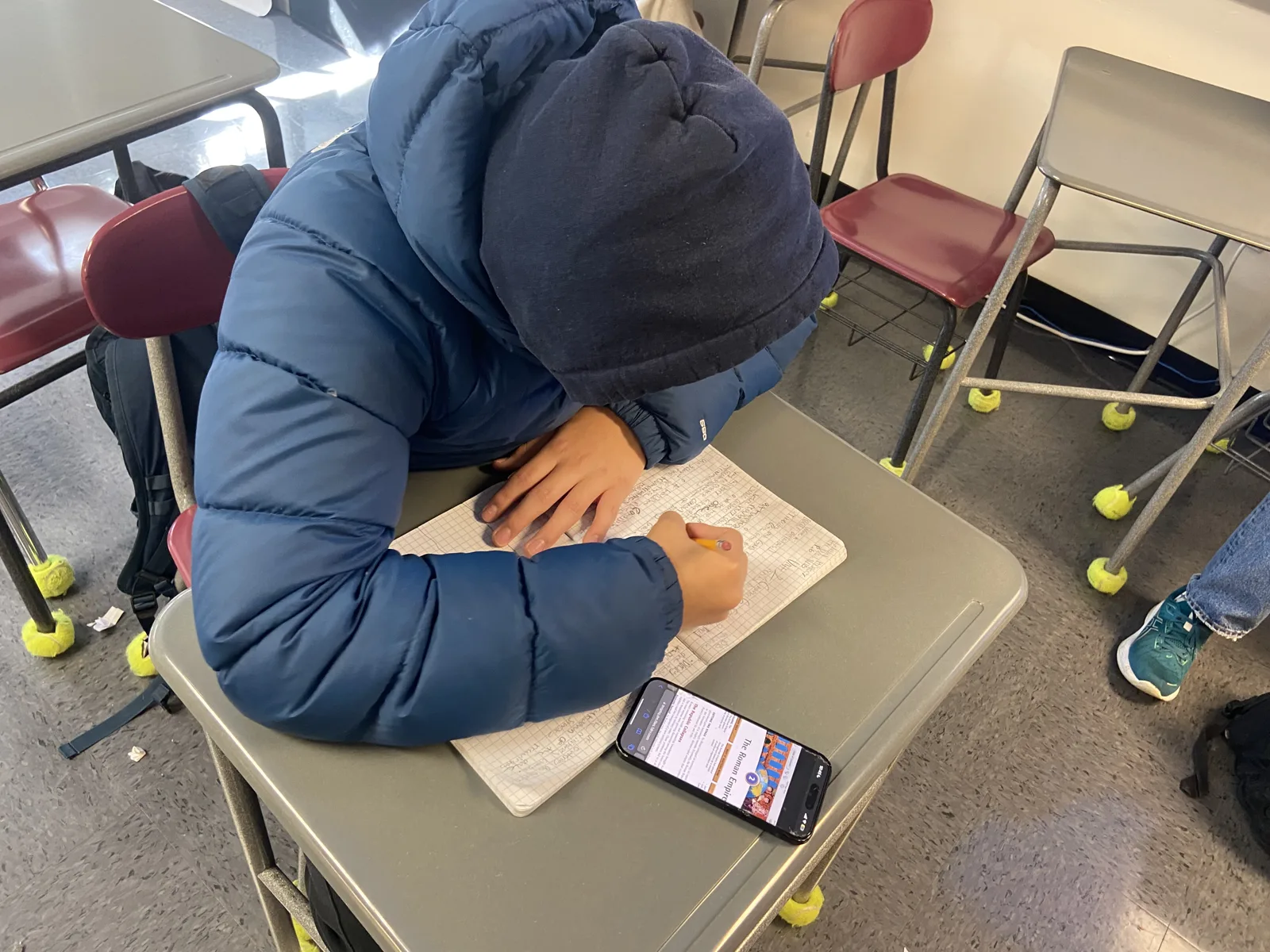 A student in a hooded blue puffer jacket bends over a notebook at a classroom desk, writing with a pencil next to a phone, with chairs and desks around